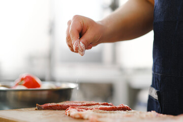 The chef salts and peppers a piece of meat. Cooking in the kitchen of restaurant.