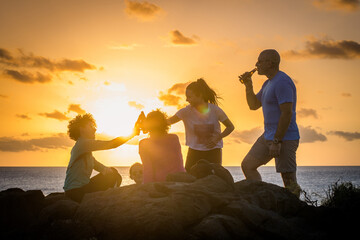 Friends toasting with bottles of beer, beautiful sunset on the ocean in background. Modern family cheering with drinks, amazing orange sky with clouds over the sea. Group of people enjoying together