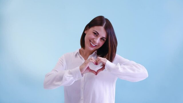Beautiful caucasian woman in white shirt smiles and shows heart symbol using her hands. Blue background. 4K resolution video. Positive emotion theme.