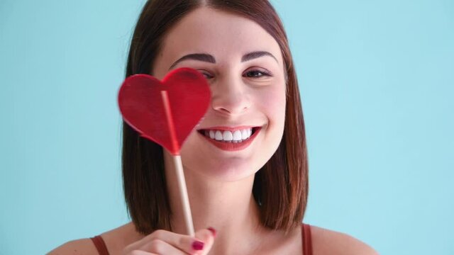 Beautiful caucasian woman with white teeth smile dances holding red heart shaped lollipop and periodically licks it. Close up portrait. 4k resolution video. Valentine's day theme.