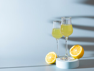 Limoncello, traditional Italian liquor on a light concrete background in the rays of the sun. Next to it is a yellow lemon, fresh citrus fruits. Composition podium.