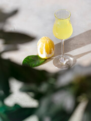 Limoncello, traditional Italian liquor on a light concrete background in the rays of the sun. Next to it is a yellow lemon, fresh citrus fruits.