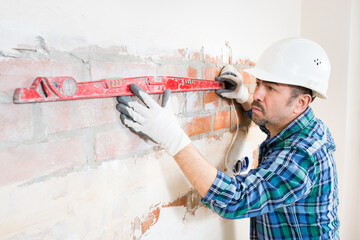 A builder in a safety helmet checks a flat brick wall with a building level while repairing a...