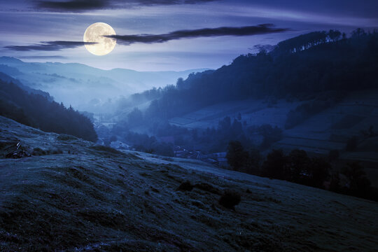 valley on the foggy night. village in the distance. grass and flowers on the hill in full moon light. beautiful countryside scenery. dark clouds on the sky