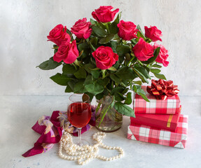A bouquet of gorgeous red roses in a vase on a light gray background. Gift boxes. Wine glass, beads, silk scarf.