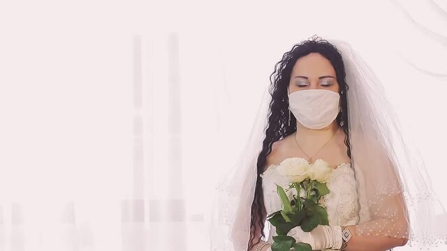 A Jewish bride in a wedding dress and a veil in a medical mask looks at the camera with smiling eyes. Medium plan
