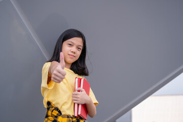 Smiling asian kid girl with holding books with showing thumbs up, education concepts