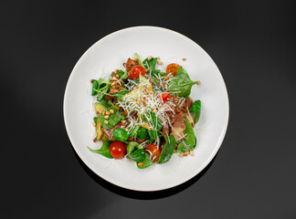 A white plate of delicious fresh salad with balsamic looks presentable. Salad of spinach, tomatoes, grilled bacon, cedar nuts, avocado and parmesan are on a black glass table top view.