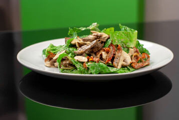 A plate with lettuce, mushrooms, grilled meats, sun-dried tomatoes and white sauce on a black glass table and green wall looks presentable. Tasty fresh salad on a white plate side vie