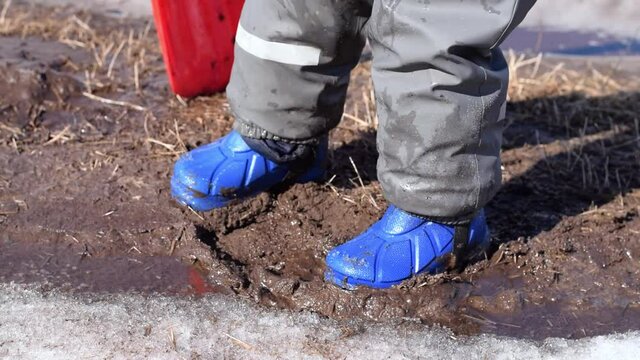 Little boy in blue rubber boots and waterproof pants is trampling in the spring mud.Spring concept.Children's leisure.Close-up, slow motion.