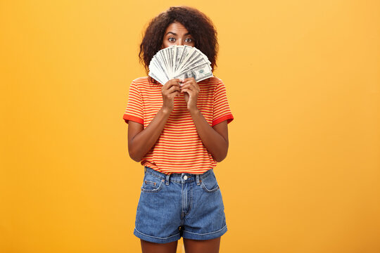 Woman gonna waste lots of cash today. Pleased and delighted rich african american girl with curly hairstyle hiding face behind bunch of money peeking mysteriously at camera winning millions
