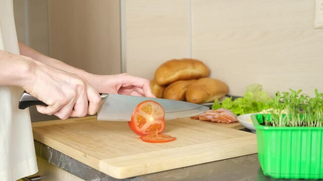 Women's hands cut tomatoes for making sandwiches on a wooden cutting board. There are other ingredients around, bread, cheese, greens. The concept of making simple meals at hds cut tomatoes