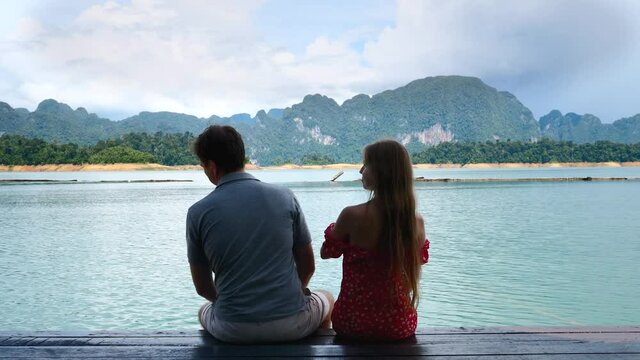 Rear View of Couple Sitting Together on Date on River Bridge, Man Flirting, Touching Woman Booty and Getting Funny Slap, Fun and Laughing Together. Pair of Travel People at Lake with Mountains