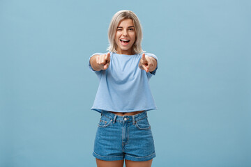 Happy enthusiastic stylish blonde female with tattoos pointing at camera as if picking or making...
