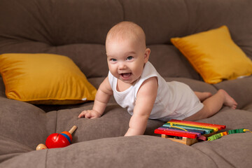 Obraz na płótnie Canvas Cute toddler in white bodysuit lies at home on grey sofa with yellow pillows playing with wooden developing toy