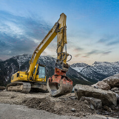 yellow chain excavator with grapple of natural stone in front of a mountain backdrop and cloudy sky