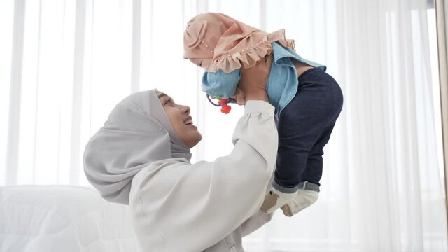 Happy asian family Mother and baby girl 6 months old in traditional hijab clothing Baby girl flying in mother's arms and kiss in white sunny bedroom. slow motion