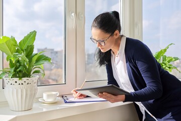 Business woman working near window using digital tablet, writing in papers