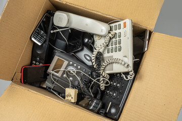 Old desk phones, cordless phone, cell phones, and smartphones in a cardboard box.