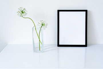 Blank black frame mockup. Small fresh flowers of white garlic (allium neapolitanum) in glass vase on glossy white table. White background, minimal and elegant space