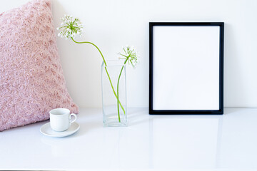 Blank black frame mockup. Small fresh flowers of white garlic (allium neapolitanum) in glass vase. Cup of coffee, pink embroidered pillow on glossy white table. Minimal and elegant work space