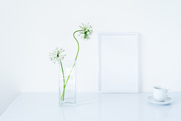Blank white frame mockup. Small fresh flowers of white garlic (allium neapolitanum) in glass vase on glossy white table. White background, minimal and elegant space