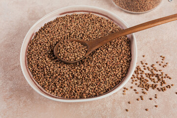Organic uncooked scattered buckwheat grain in a bowl with wooden spoon on a light beige background