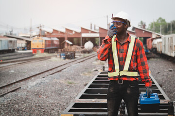 African machine engineer technician talking with walkie talkie and wearing a helmet, groves and safety vest is using a wrench to repair the train
