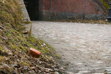 A close up on a single red brick laying next to a grass covered hill located next to a stone covered path leading to a red brick gate seen on a sunny spring day on a Polish countryside during a hike