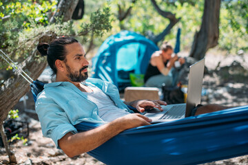 A man works on a laptop while lying in a hammock.