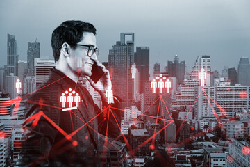 A young handsome eastern businessman processing conference call to develop social media marketing strategy to achieve business goals. Hologram icons over Bangkok background.