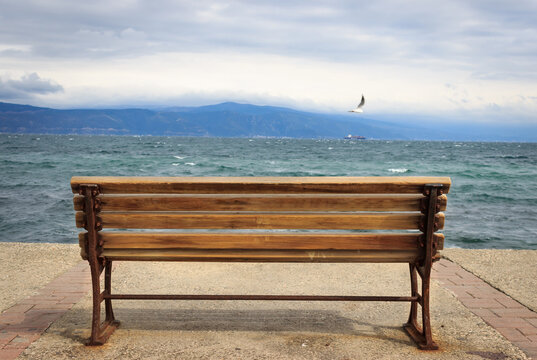 An empty wooden bench near the sea. Blue sea and mountain landscape at the viewpoint of bench. Bird is flying in cloudy day.