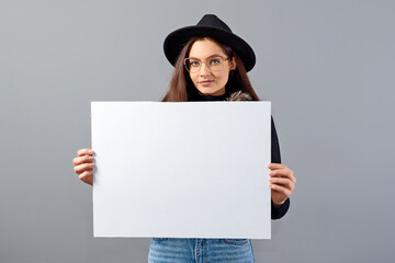 elegant young woman in glasses and hat showing empty board banner, isolated on gray, copy space