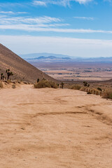 Fototapeta na wymiar Sandy dirt road leading by mountainside towards desert valley below under a cloudy sky.