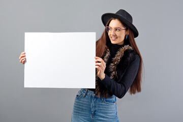 elegant young woman in glasses and hat showing empty board banner, isolated on gray, copy space