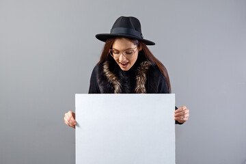 elegant young woman in glasses and hat showing empty board banner, isolated on gray, copy space