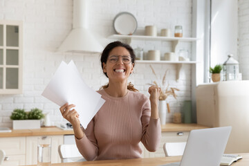 Overjoyed young latina woman in glasses scream yes hold official paper letter in hand confirming...