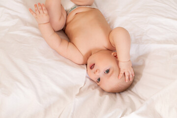 Cute baby in diapers lies in bed with white cotton linen, view from above, space for text