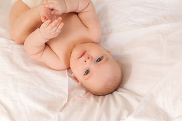 Cute baby in diapers lies in bed with white cotton linen, view from above, space for text