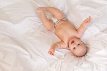 Cute baby in diapers lies in bed with white cotton linen, view from above, space for text