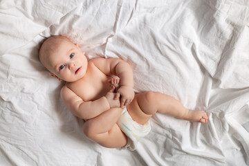 Cute baby in diapers lies in bed with white cotton linen, view from above, space for text