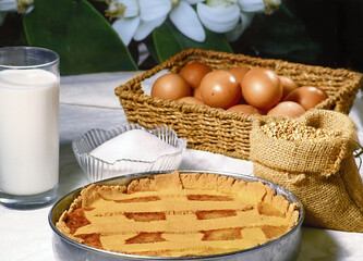 Gluten-free Neapolitan Pastiera with the ingredients in a cup full of sugar, a basket of fresh eggs, a bottle of milk and a hemp bag full of buckwheat