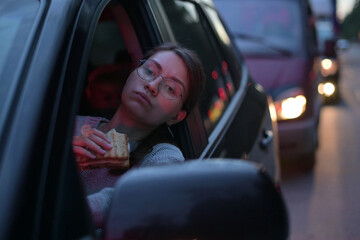 Young woman eating a sandwich while standing in a traffic jam. Food on the go, rush hour