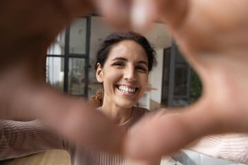 Cheerful affectionate hispanic woman take romantic selfie portrait looking at camera through heart of joined fingers. Enamored young lady shoot love confession to beloved man on video at Valentine day