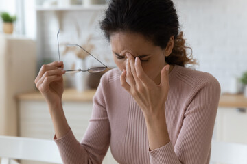 Having problem with eyes. Sad fatigue millennial latina female sit at home kitchen take off...