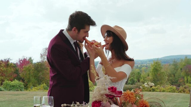 Stylish young bride and groom feed each other pizza at wedding reception, Slowmo