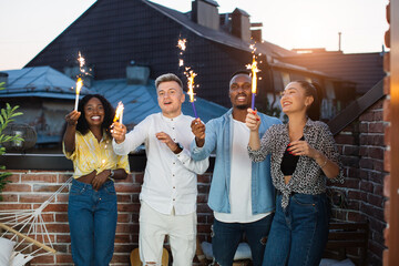 Group of four cheerful friends in stylish outfits celebrating party on roof with sparklers. Young diverse people hanging out together during weekends.