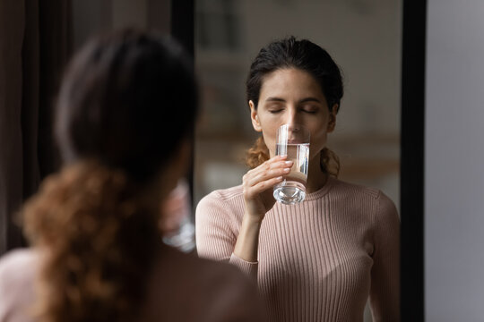 Mirror reflection of young hispanic lady drink fresh clear tasty natural mineral water from glass enjoy has pleasure. Pretty female hydrate sip purified still aqua at morning take care of skin beauty