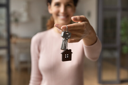 Please take your keys. Close up blurred portrait of smiling young latin lady real estate owner seller propose you client to get key from new home flat. Focus on female broker hand giving bunch of keys