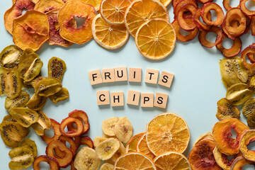 Dried sliced plums, kiwi, peach, orange fruits chips on blue background and text - Fruits chips. Snack vegan free sugar food. View from above.